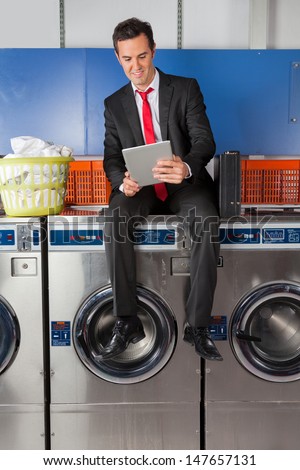 Full length of young businessman using digital tablet while sitting on washing machine in laundry