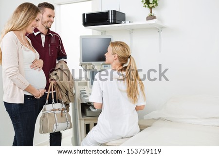 Young female doctor looking at expectant couple while using ultrasound machine in clinic
