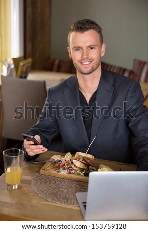 Portrait of smart businessman with mobilephone and laptop having meal in restaurant