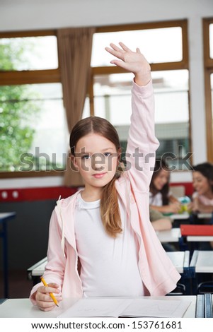 Portrait of cute schoolgirl raising hand while standing at desk in classroom