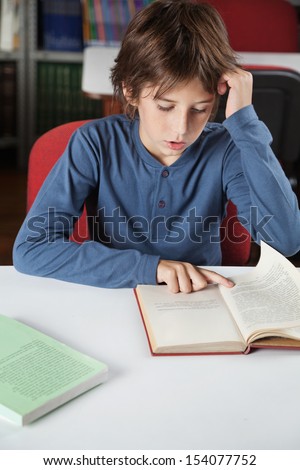 Little schoolboy reading book at table in library