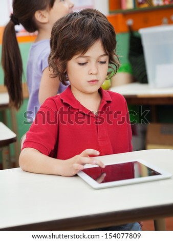 Little boy using digital at desk with friend in background at kindergarten