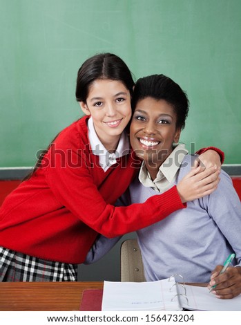 Portrait of little schoolgirl hugging female teacher at desk in classroom