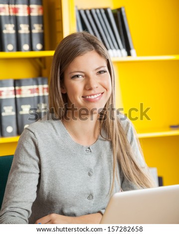 Portrait of beautiful female student with laptop smiling against bookshelf in college library