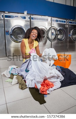Happy young African American woman sitting with clothes on floor at laundry