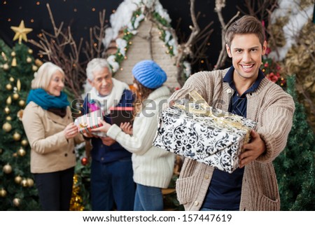 Portrait of happy man offering Christmas present with family standing in background at store