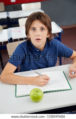 Portrait of shocked little boy with cheat sheet sitting at desk in classroom
