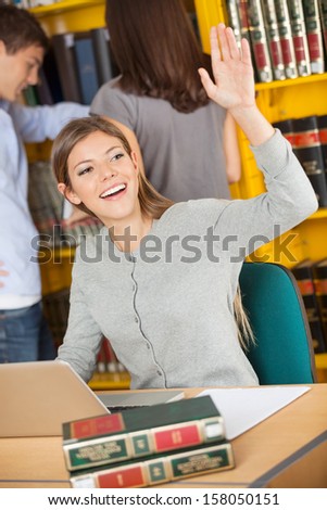 Happy female student with laptop and books looking away while waving in college library