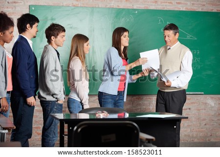 Mature male teacher giving test result to student while classmates standing in row at classroom