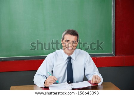 Portrait of happy male teacher with binder and pen sitting at desk in classroom