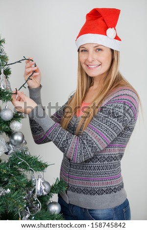 Portrait of beautiful woman in Santa hat decorating Christmas tree with fairy lights at home
