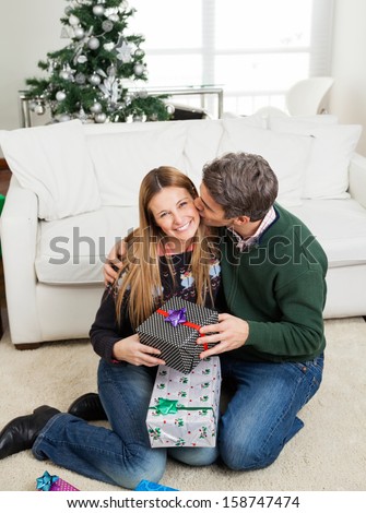 Loving mid adult man with Christmas gift kissing woman on cheek at home