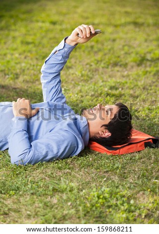 Side view of young male student reading text message on mobilephone while lying on grass at college campus