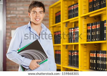 Portrait of confident male student holding books while standing in university campus