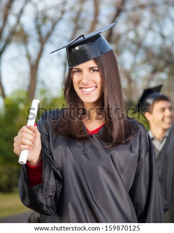 Portrait of happy young woman in graduation gown holding diploma on college campus