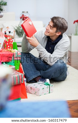 Full length of happy man looking at Christmas present while sitting on floor in house