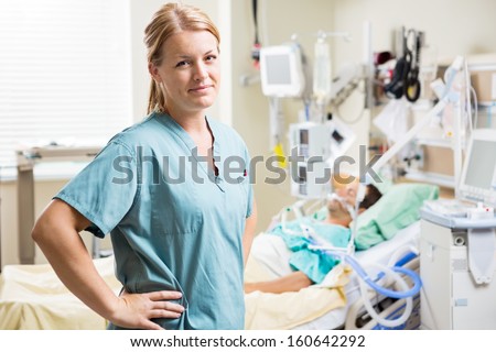 Portrait of confident nurse standing with hand on hip while patient resting in background at hospital