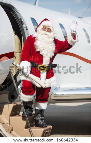 Full length portrait of Santa waving hand while standing on private jet\'s ladder at airport terminal
