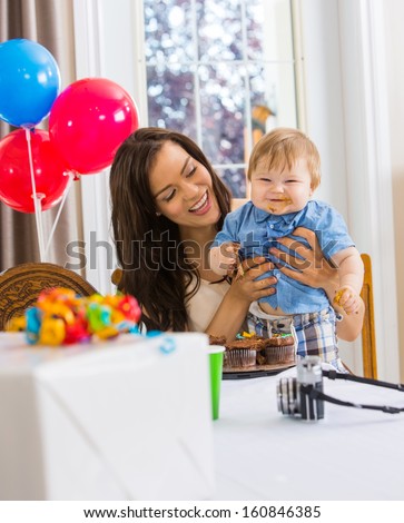 Happy mother holding baby boy with messy hands covered with cake icing at birthday party