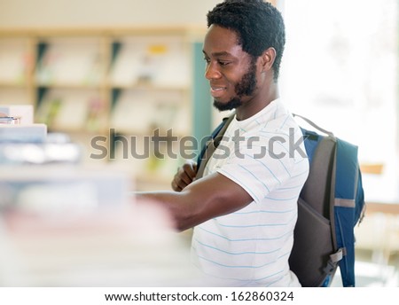 Side view of young student carrying backpack while choosing book at library
