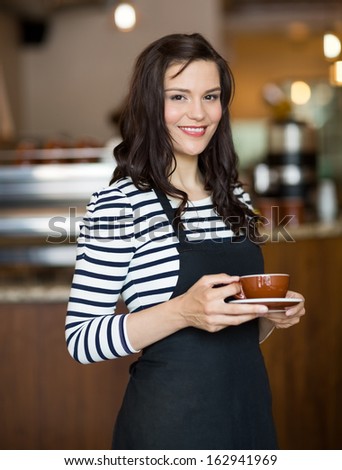 Portrait of attractive waitress holding coffee cup while standing in cafeteria