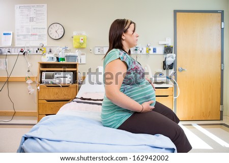 Side view of thoughtful pregnant woman with hands on stomach sitting on hospital bed