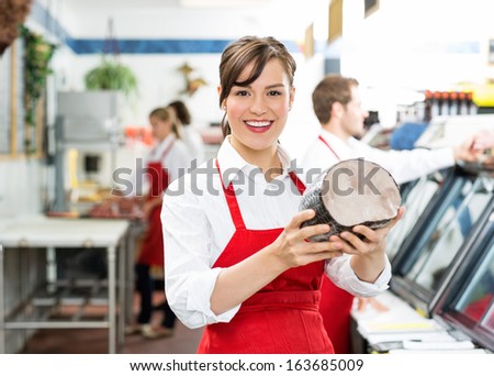 Portrait of happy female butcher holding ham at store with colleagues working in background