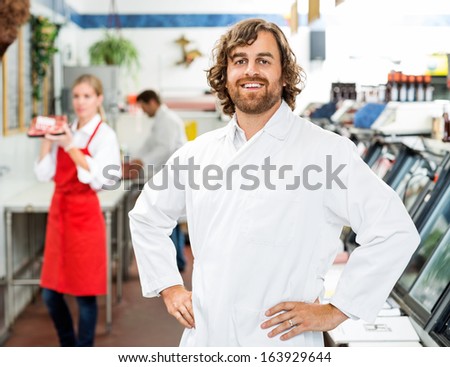 Portrait of confident butcher standing at store with colleagues working in background