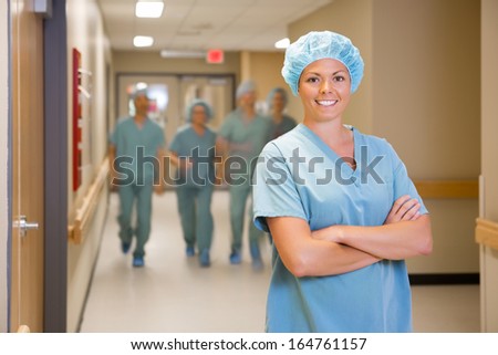 Portrait of happy female surgeon standing arms crossed with team walking in hospital corridor