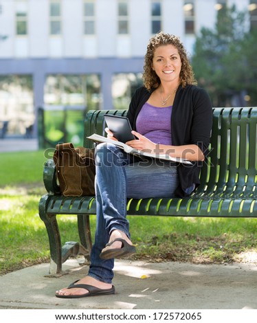 Portrait of smiling female student with book and digital tablet sitting on bench at university campus