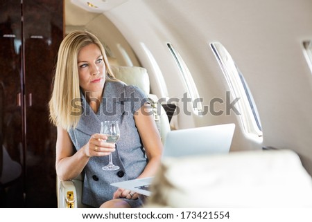 Beautiful businesswoman holding wineglass while looking through window of private jet