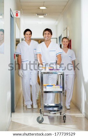 Portrait of confident lab technicians with medical cart walking in hospital corridor