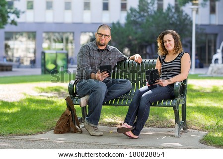 Portrait of confident students with digital tablets sitting together on bench at university campus