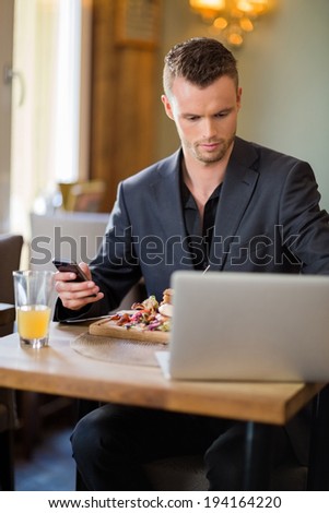 Young businessman with cellphone and laptop having sandwich in restaurant