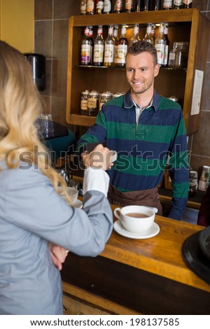 Young male bartender holding card-reader while woman making payment through cellphone at coffeeshop