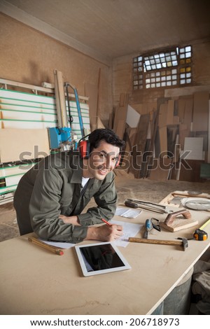 Portrait of handsome carpenter working on blueprint while wearing ear protectors at table in workshop