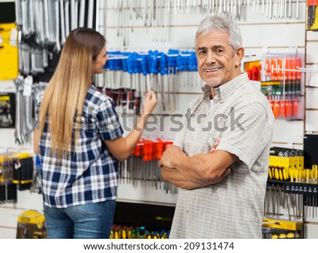Portrait of happy senior man standing arms crossed with daughter in background at hardware store
