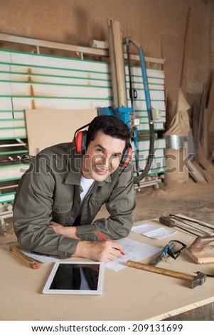Portrait of confident carpenter working on blueprint while wearing ear protectors at table in workshop