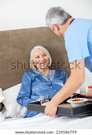 Male caretaker serving breakfast to happy senior woman on bed in nursing home