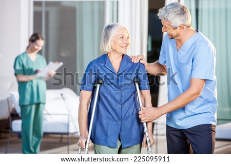 Happy male caretaker helping senior woman in walking with nurse in background at nursing home yard