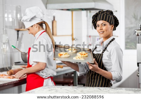 Happy female chef holding small pizzas on tray with colleague working in background at commercial kitchen