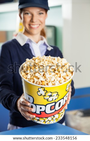 Portrait of happy female worker offering popcorn bucket at cinema concession stand