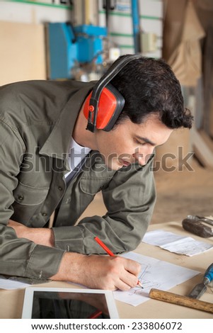 Young carpenter working on blueprint while wearing ear protectors at table in workshop