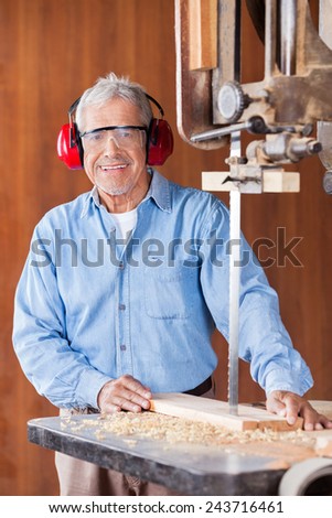 Portrait of happy senior carpenter cutting wood with bandsaw in workshop
