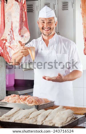 Portrait of happy mature butcher showing chicken piece at counter in shop