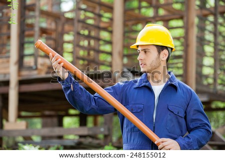 Young male worker examining pipe against incomplete timber cabin at construction site