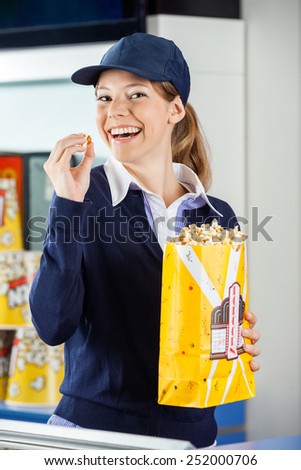 Portrait of cheerful female worker eating popcorn from paperbag at cinema concession stand