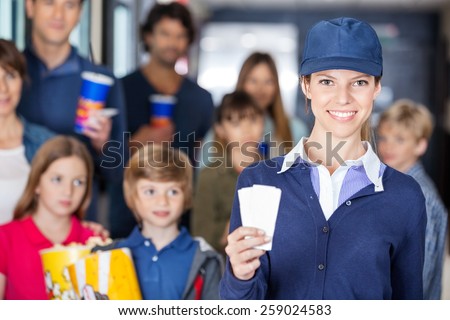 Portrait of confident female worker holding tickets while families waiting in background at cinema