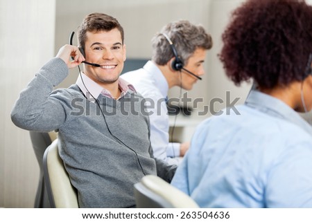 Portrait of young smiling male customer service representative using headset while colleagues working in office