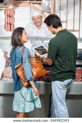Mature couple using digital tablet while standing at counter of butcher shop